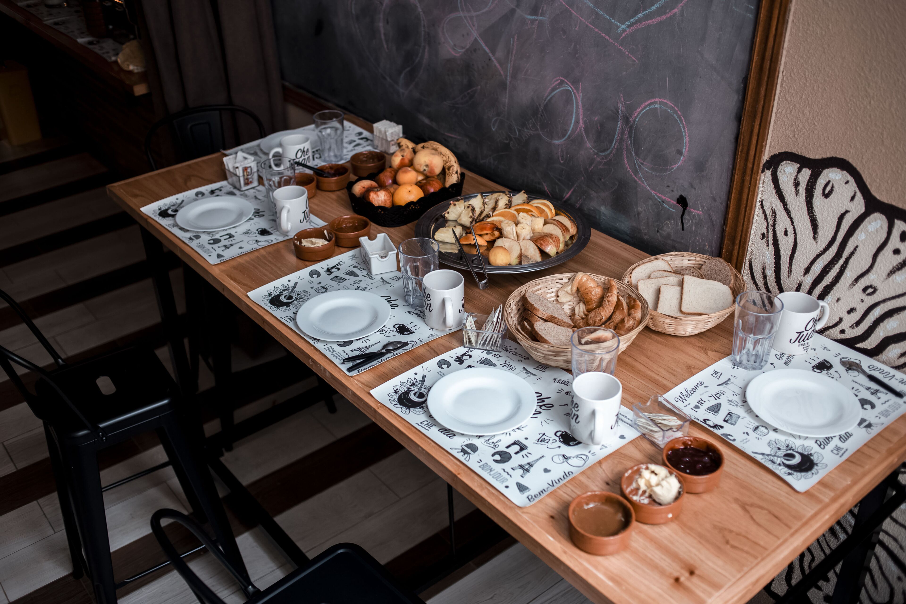A dining table set with plates, cups, and various food items, including bread and pastries, in a room with a chalkboard wall.