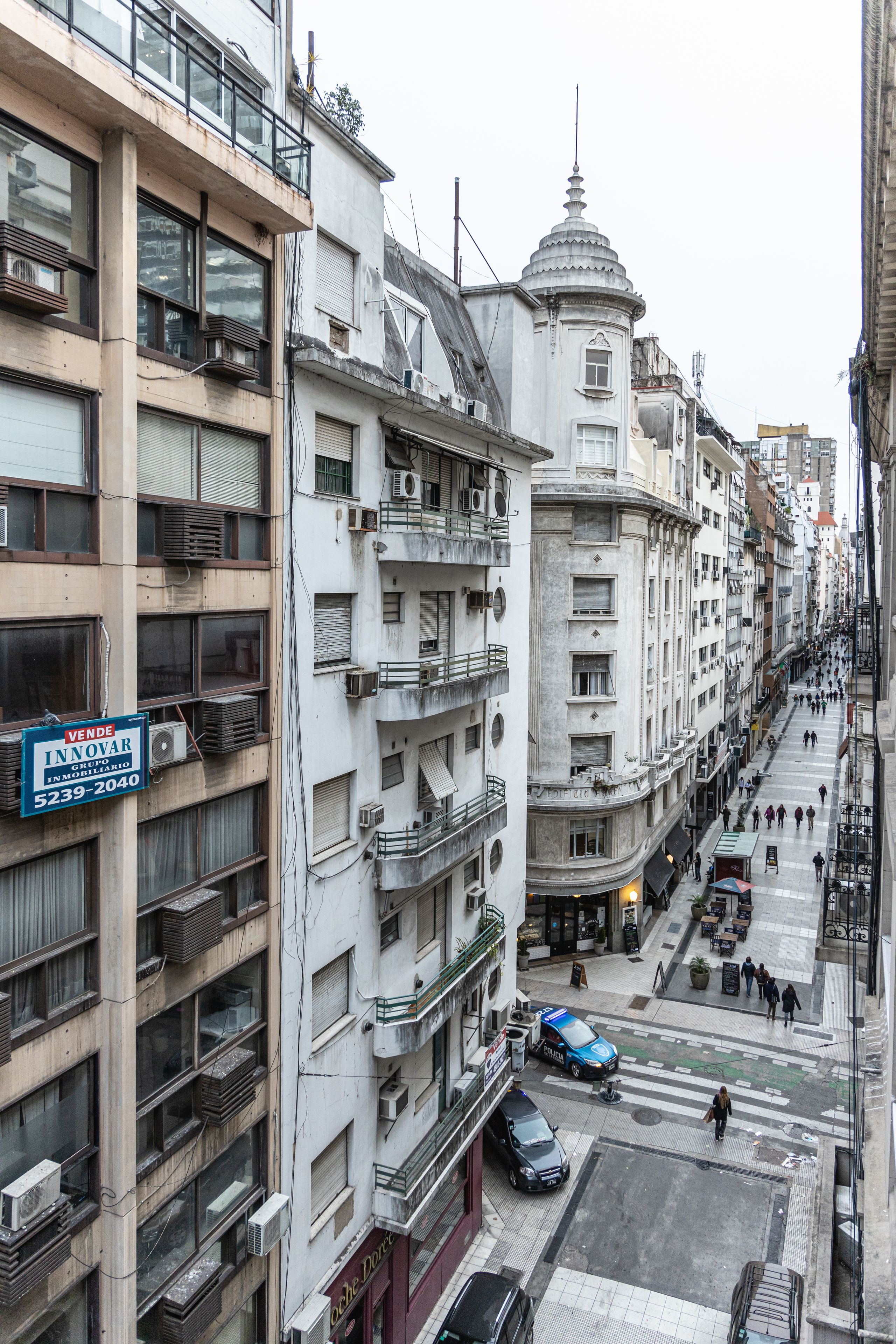 A multi-story building with balconies, a dome structure, and a street with pedestrians and parked cars.