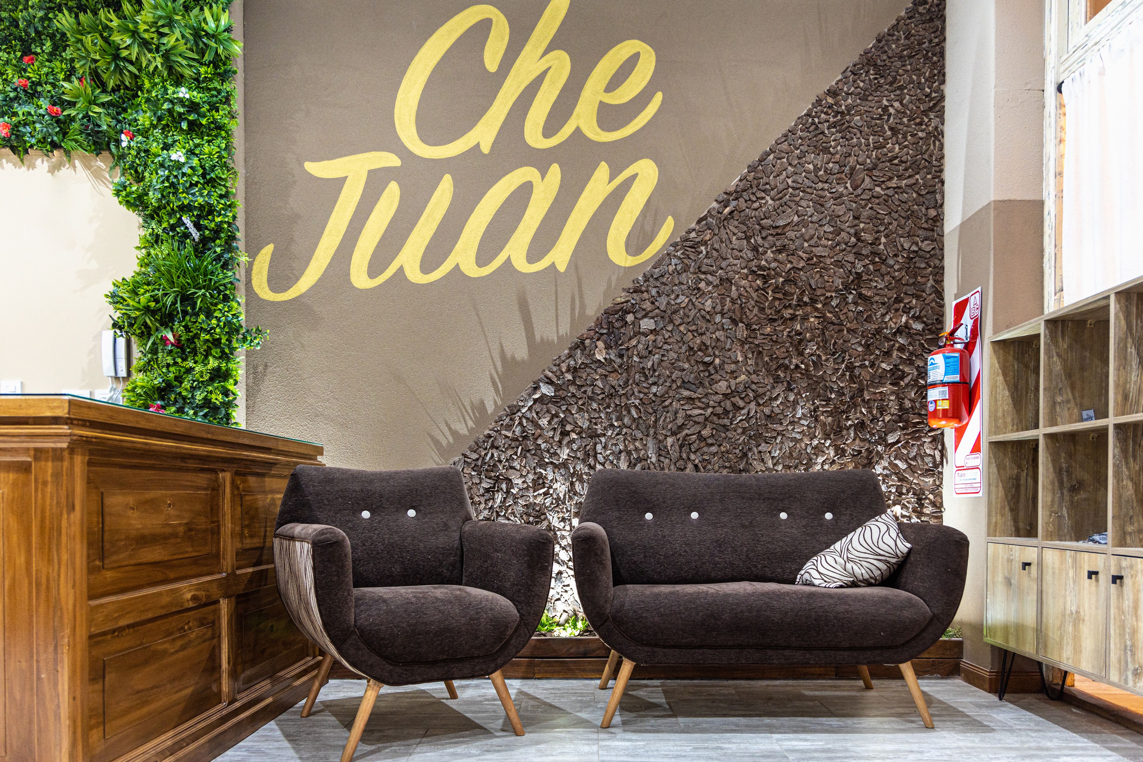 A lobby with two dark armchairs, a wooden reception desk, and a large wall with 'Che Juan' written on it.