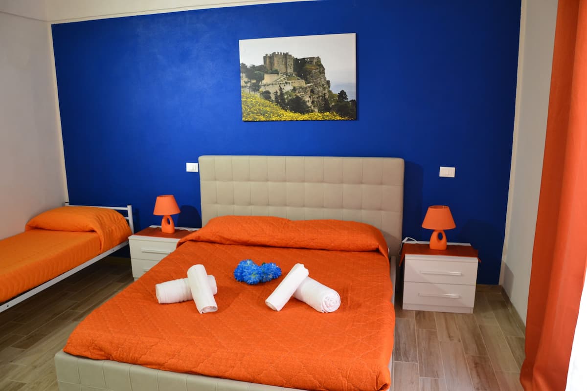 Habitación de hotel con dos camas, ropa de cama naranja, una cama grande con toallas enrolladas, una mesita de noche con lámpara y un cuadro de un castillo en la pared.