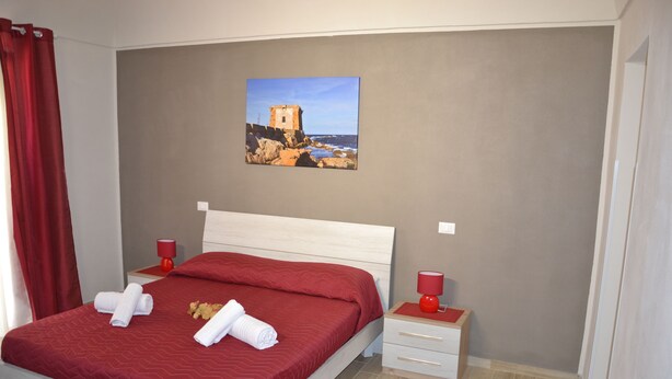 A neatly made bed with red bedding, two bedside tables with red lamps, and a painting of a coastal scene on the wall. - B&B Stella Maris Trapani Centro (Trapani)
