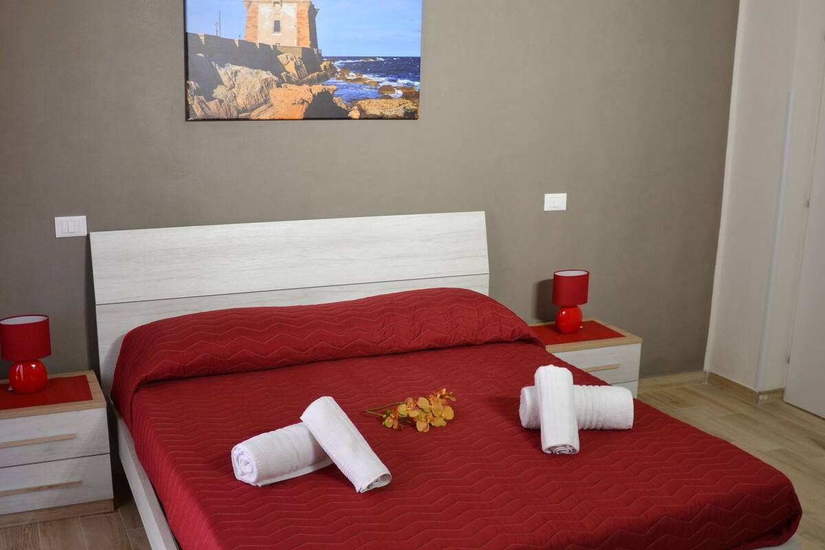 Una cama bien hecha con ropa de cama roja, dos toallas enrolladas y un pequeño ramo de flores.