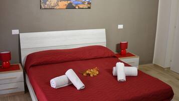 A neatly made bed with red bedding, two rolled towels, and a small bouquet of flowers.