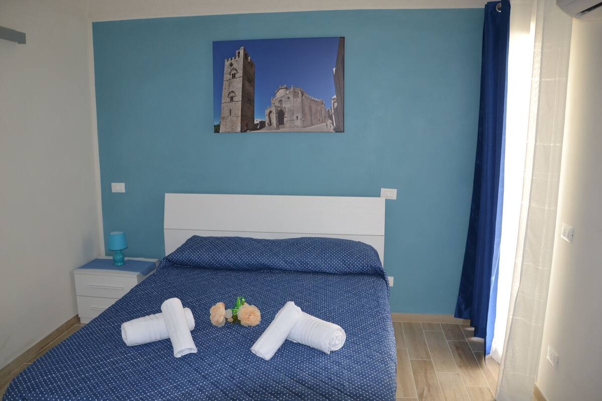 Una cama bien tendida con ropa de cama azul, una mesita de noche pequeña con una lámpara y un cuadro enmarcado de un edificio histórico en la pared.