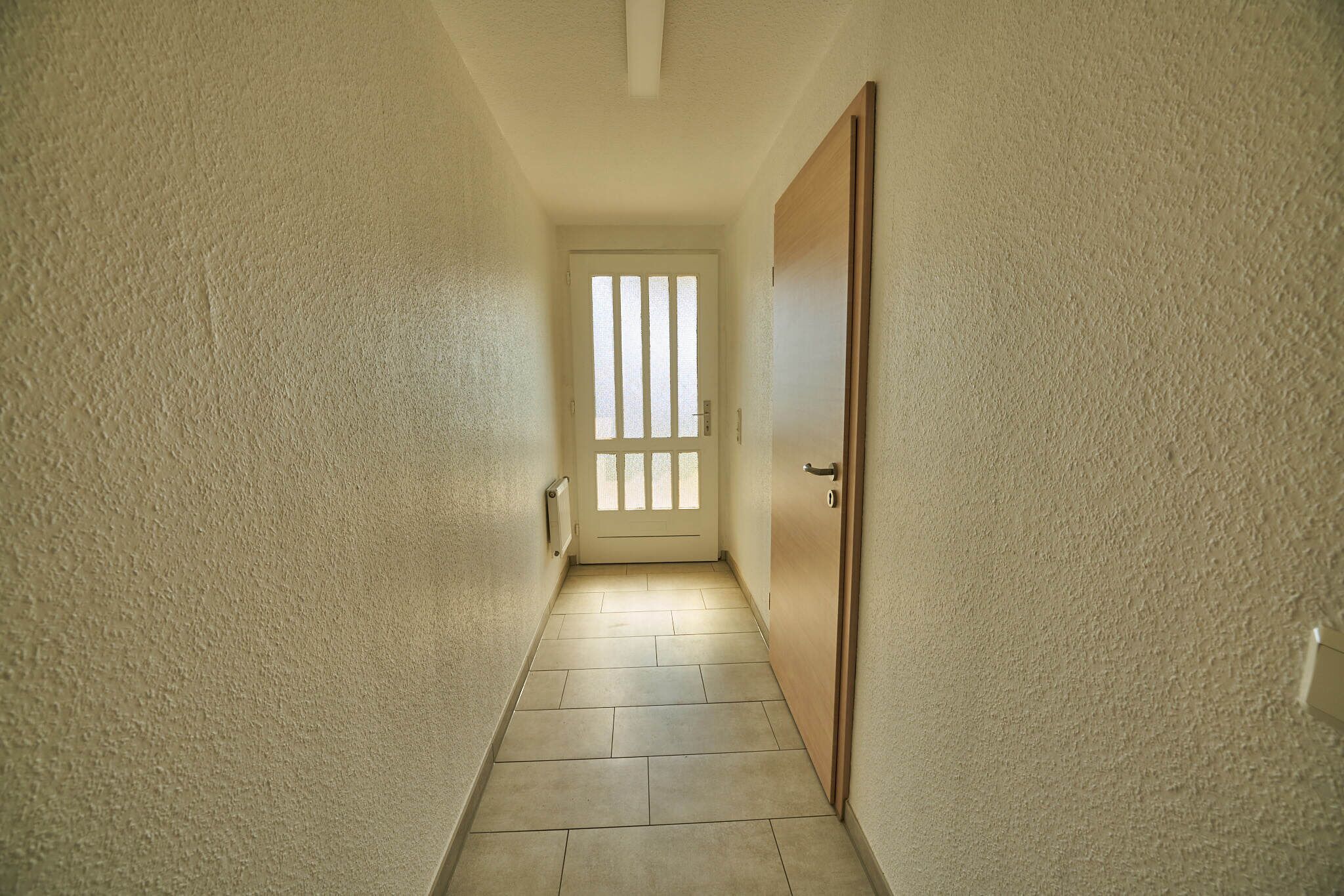 Un pasillo estrecho con una puerta de madera al final, que da a una habitación con ventana.