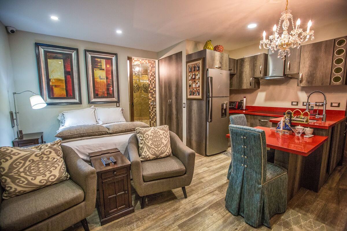 A modern kitchen with a refrigerator, stove, and sink. There is a dining area with a red table and chairs. The room has a bed with a white comforter and two decorative pillows.