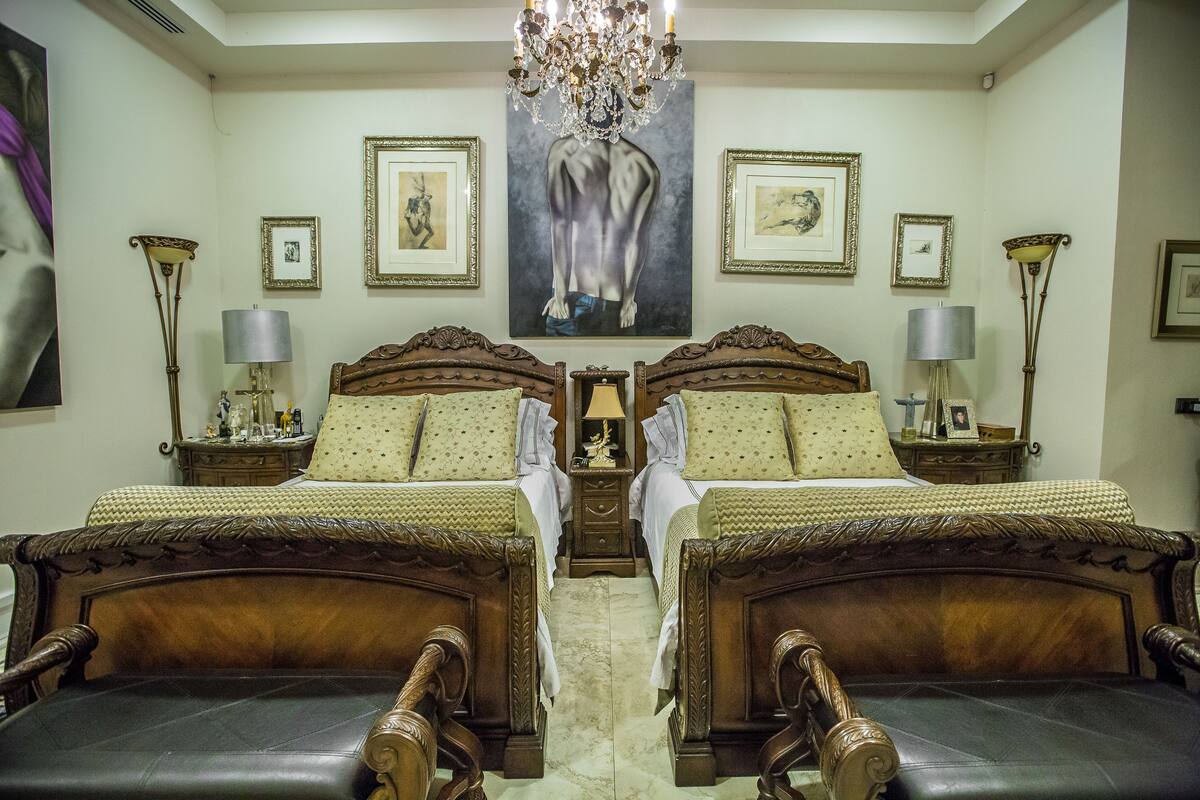 A hotel room with two wooden beds, a chandelier, framed artwork on the walls, and bedside tables with lamps.
