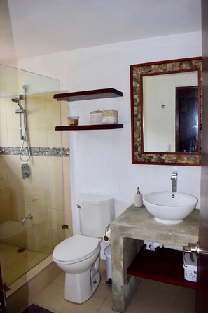 Et badeværelse med glasbrusekabine, toilet, vask og et spejl med stenkant.