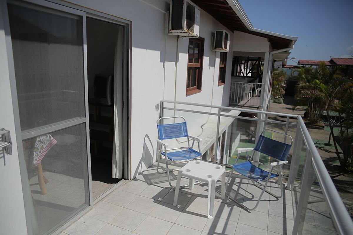 A balcony with two blue chairs and a small table, a sliding glass door, and a white building with a red roof.