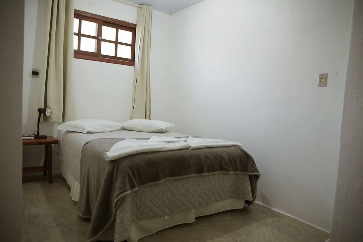 A small, clean room with a single bed, a nightstand, a window with curtains, and a light switch on the wall.