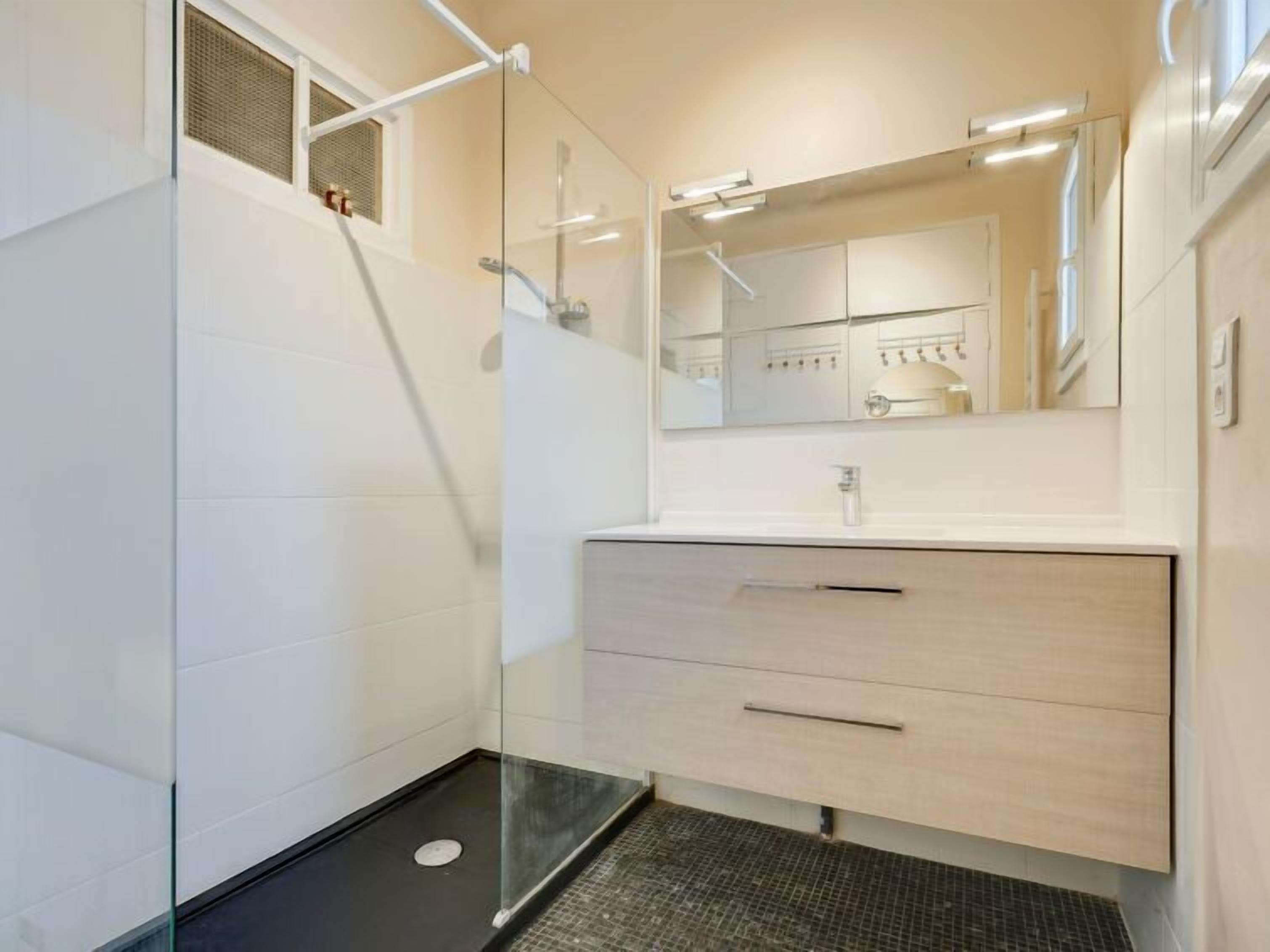 Een moderne badkamer met een douchecabine van glas, een wit badkamermeubel met spiegel, en een betegelde vloer.