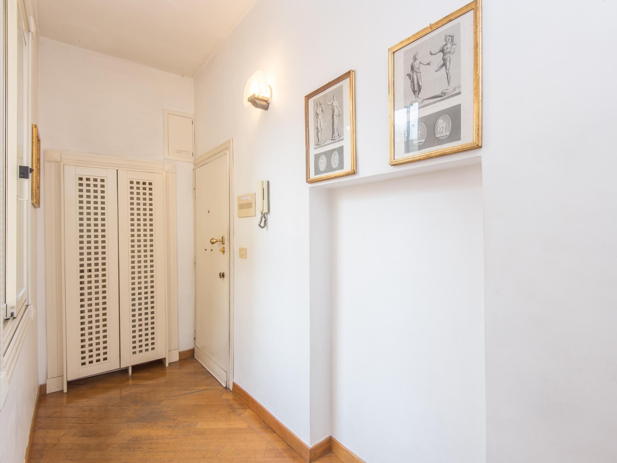 Un couloir doté d’un plancher en bois, de murs blancs et de deux portes avec des panneaux de verre à persiennes. Des tableaux encadrés ornent le mur et un luminaire est fixé au-dessus de la porte.