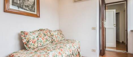 A room with a floral bedspread, a framed picture on the wall, and an air conditioning unit.