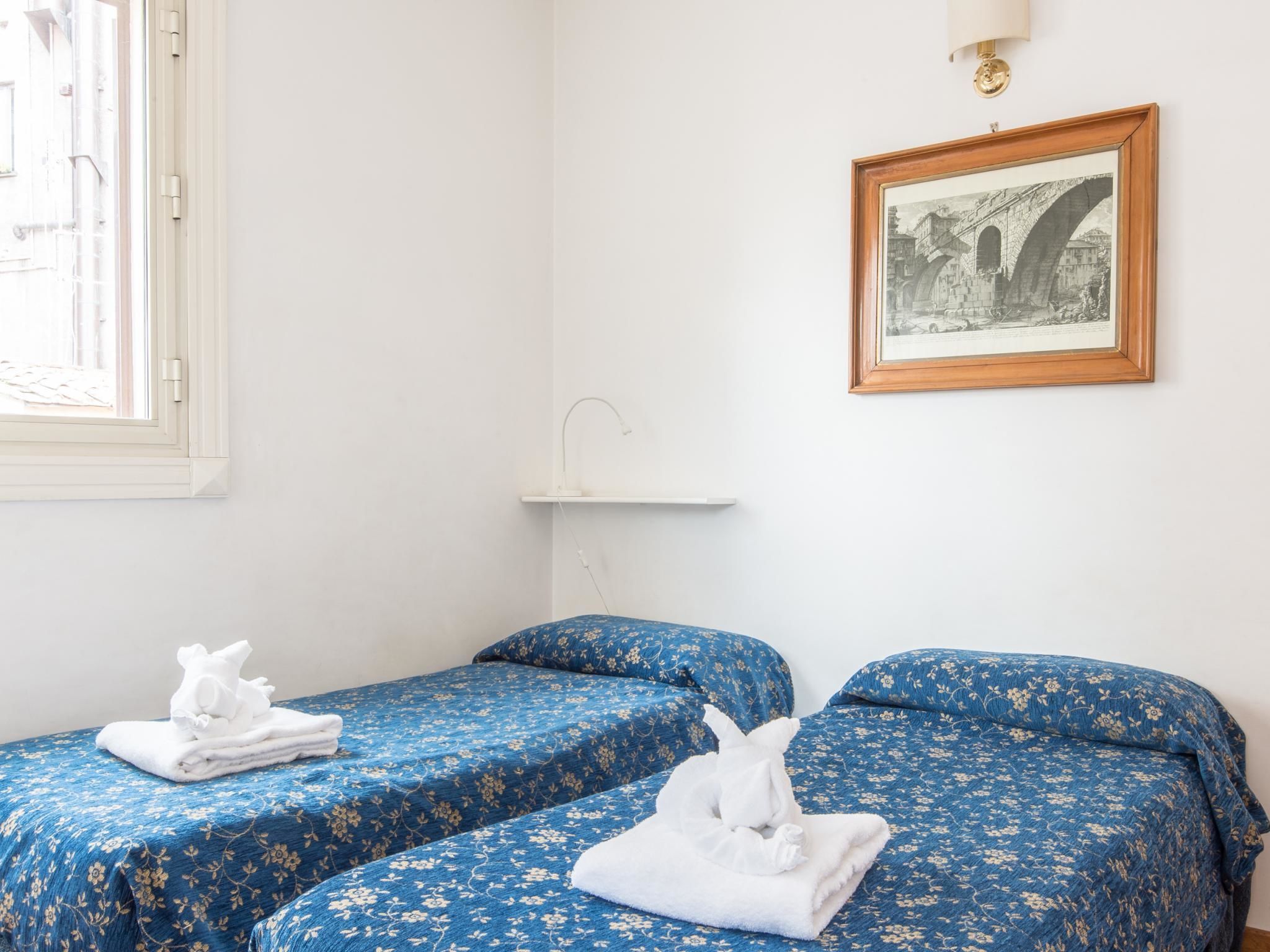 A hotel room with two beds, each with a blue floral bedspread and white towels. There is a framed picture on the wall and a window with a view of the outside.