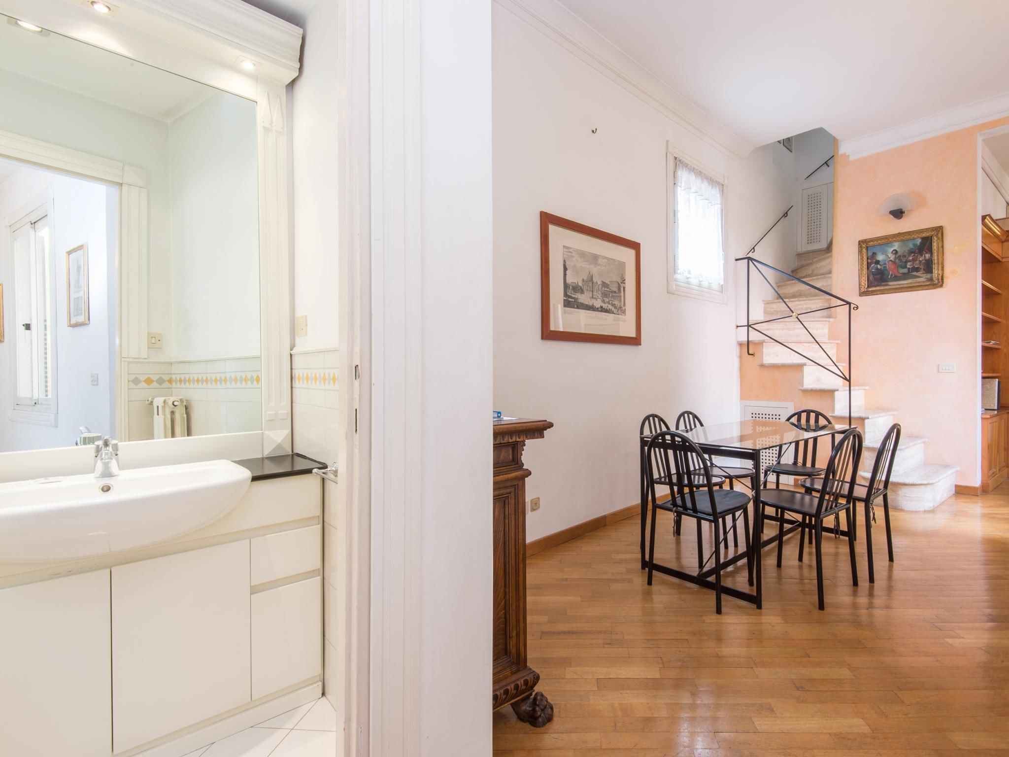 A spacious room with a large white sink, wooden dining table with chairs, a staircase, and a framed picture on the wall.