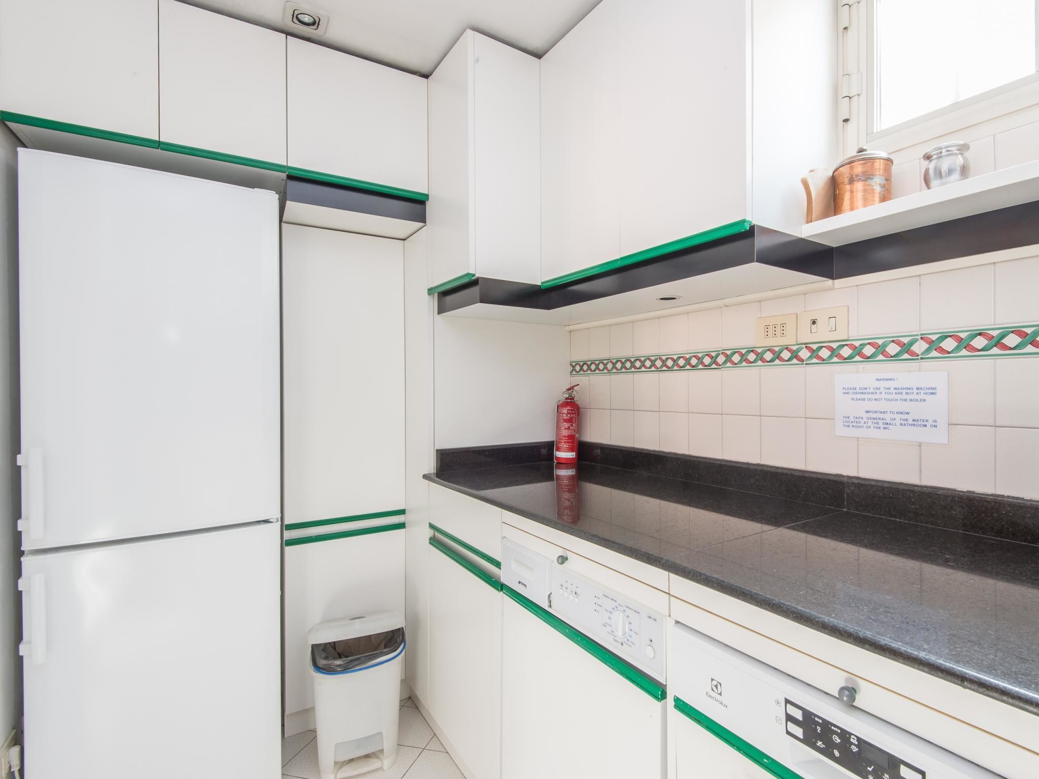 Una cocina compacta con armarios blancos, detalles en verde, una encimera negra y un extintor a la vista.