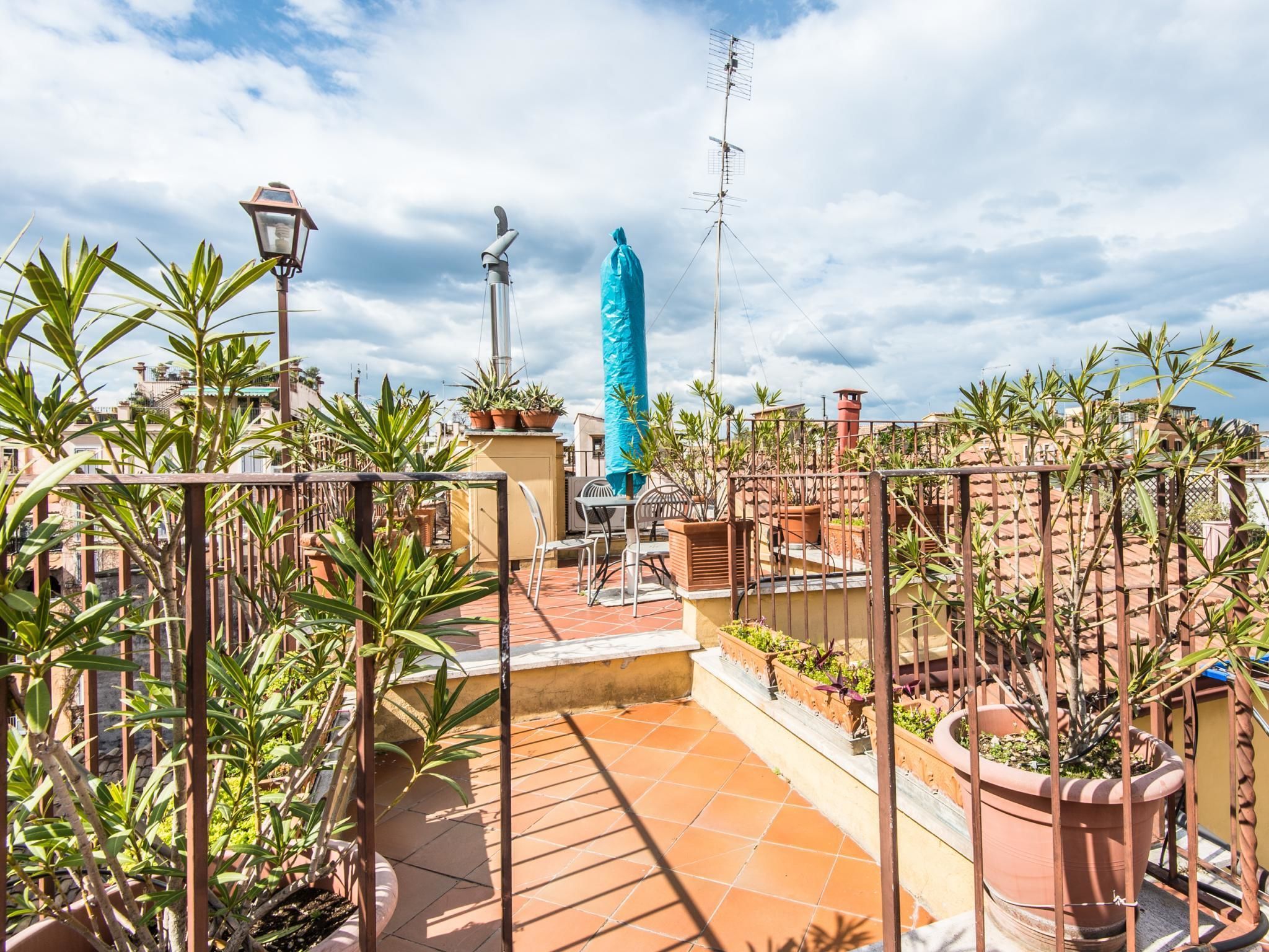 A rooftop terrace with potted plants, a blue umbrella, and a metal fence.