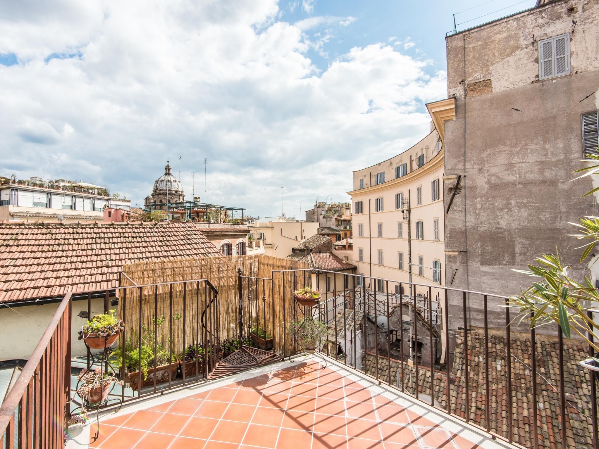 A rooftop terrace with terracotta tiles, potted plants, and a view of historic buildings and a dome structure in the background.
