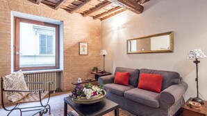 A cozy living room with a grey sofa, red cushions, a wooden coffee table with a floral centerpiece, a black metal bench, and a window with shutters. - RSH Elegant Apartment Campo dei Fiori (Rome)