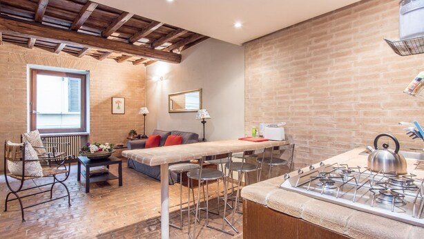 A cozy living area with a sofa, coffee table, and a dining table with chairs. There is a kitchen area with a gas stove and a brick wall. - RSH Elegant Apartment Campo dei Fiori (Rome)