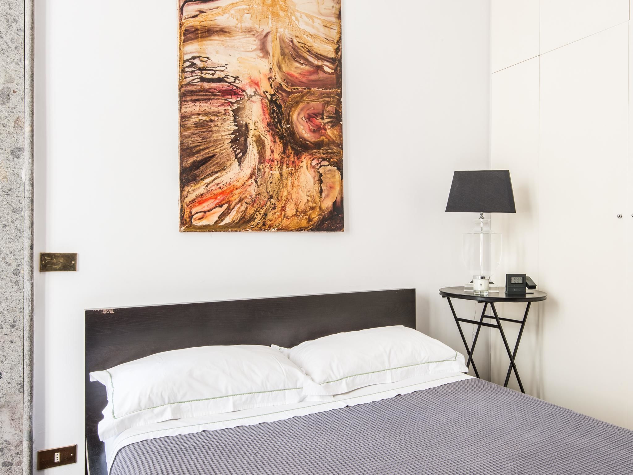 A hotel room with a bed, a bedside table with a lamp, and a large abstract painting on the wall.