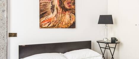 A hotel room with a bed, a bedside table with a lamp, and a large abstract painting on the wall.