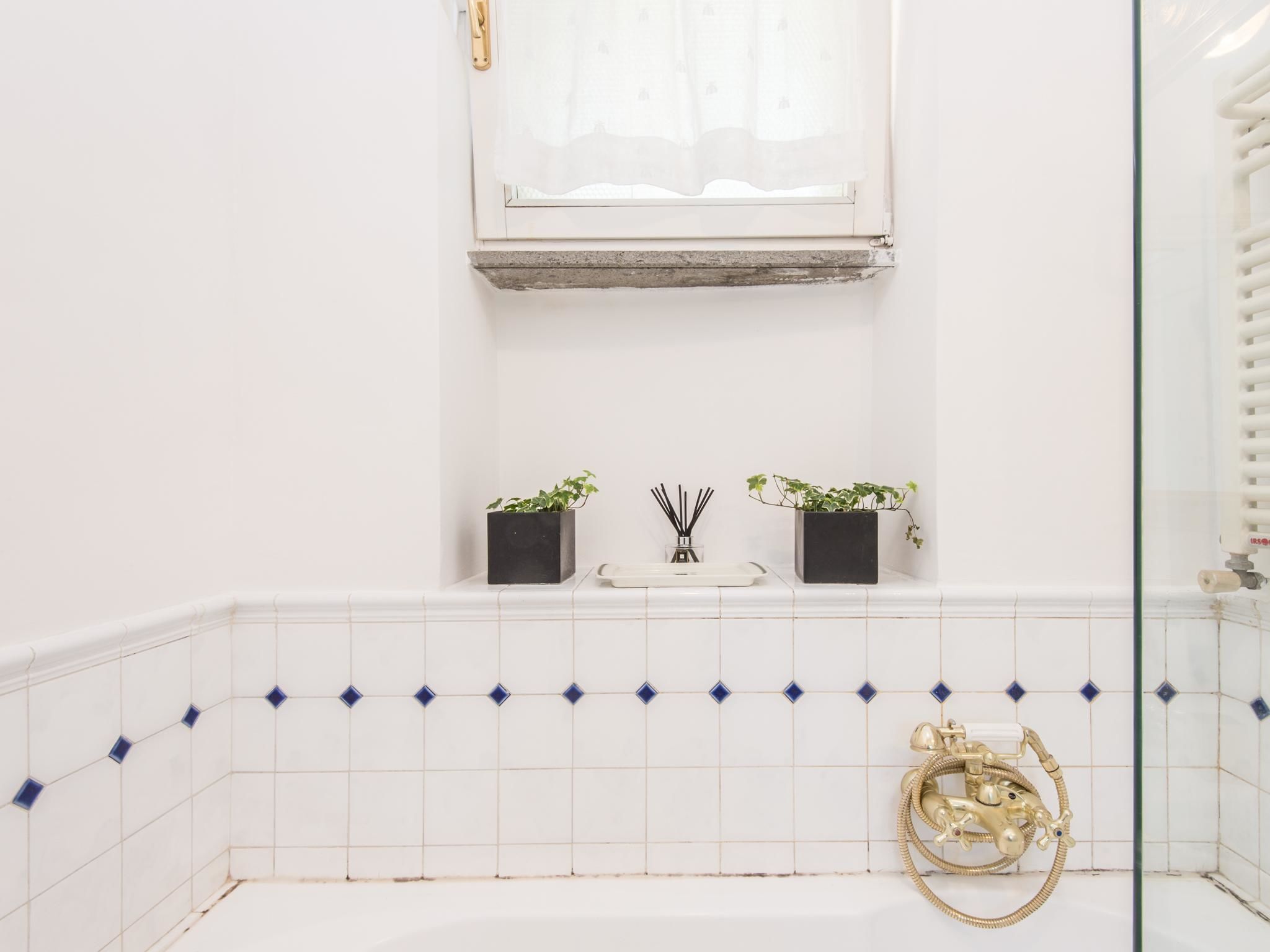 A bathroom with white tiled walls, a window with a sheer curtain, a small shelf with two potted plants, a white tray, and a gold showerhead.