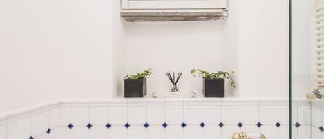 A bathroom with white tiled walls, a window with a sheer curtain, a small shelf with two potted plants, a white tray, and a gold showerhead.