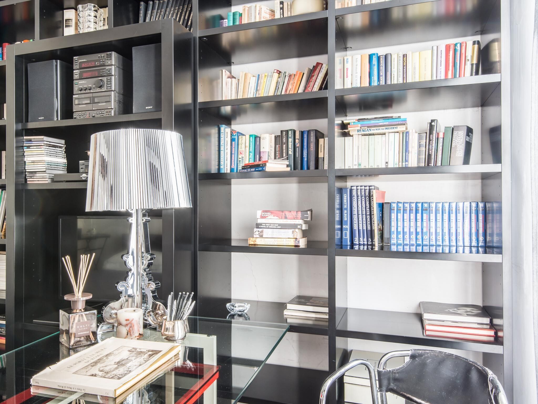 Et moderne bibliotek med sorte reoler fyldt med bøger, et glastæppe med en lampe og en stol.