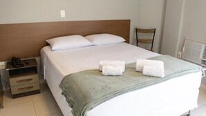 Un lit bien fait, avec du linge de lit blanc et un plaid vert, une table de chevet avec un téléphone et une télécommande, et une chaise dans un coin.