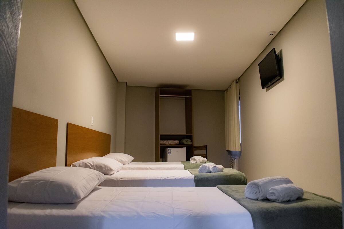 Quarto de hotel com três camas de solteiro, uma escrivaninha pequena, um espelho e uma televisão na parede.