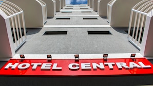 L’entrée d’un hôtel, avec une enseigne rouge portant l’inscription « HÔTEL CENTRAL » et une façade symétrique dotée de nombreuses fenêtres et de balcons.