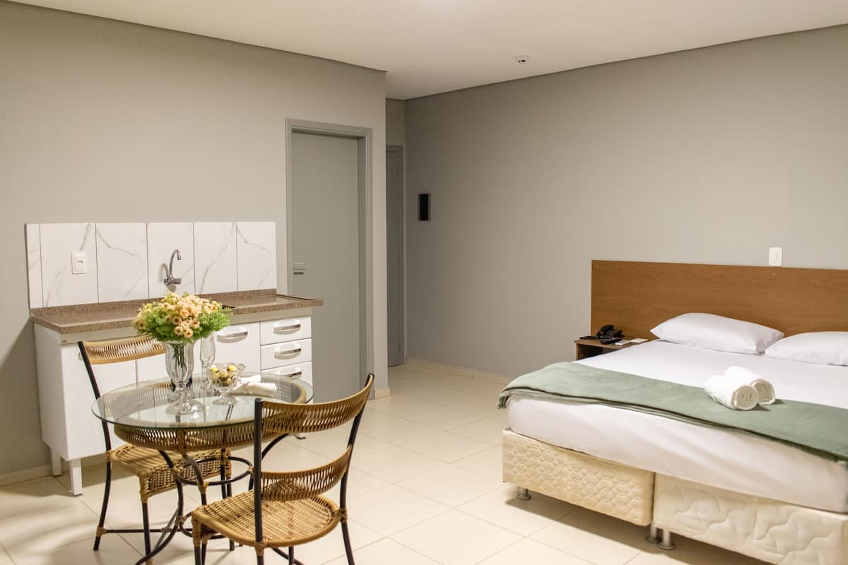 Quarto de hotel moderno com uma cama, uma pequena escrivaninha com cadeira, uma mesa de vidro com cadeiras e um vaso de flores.