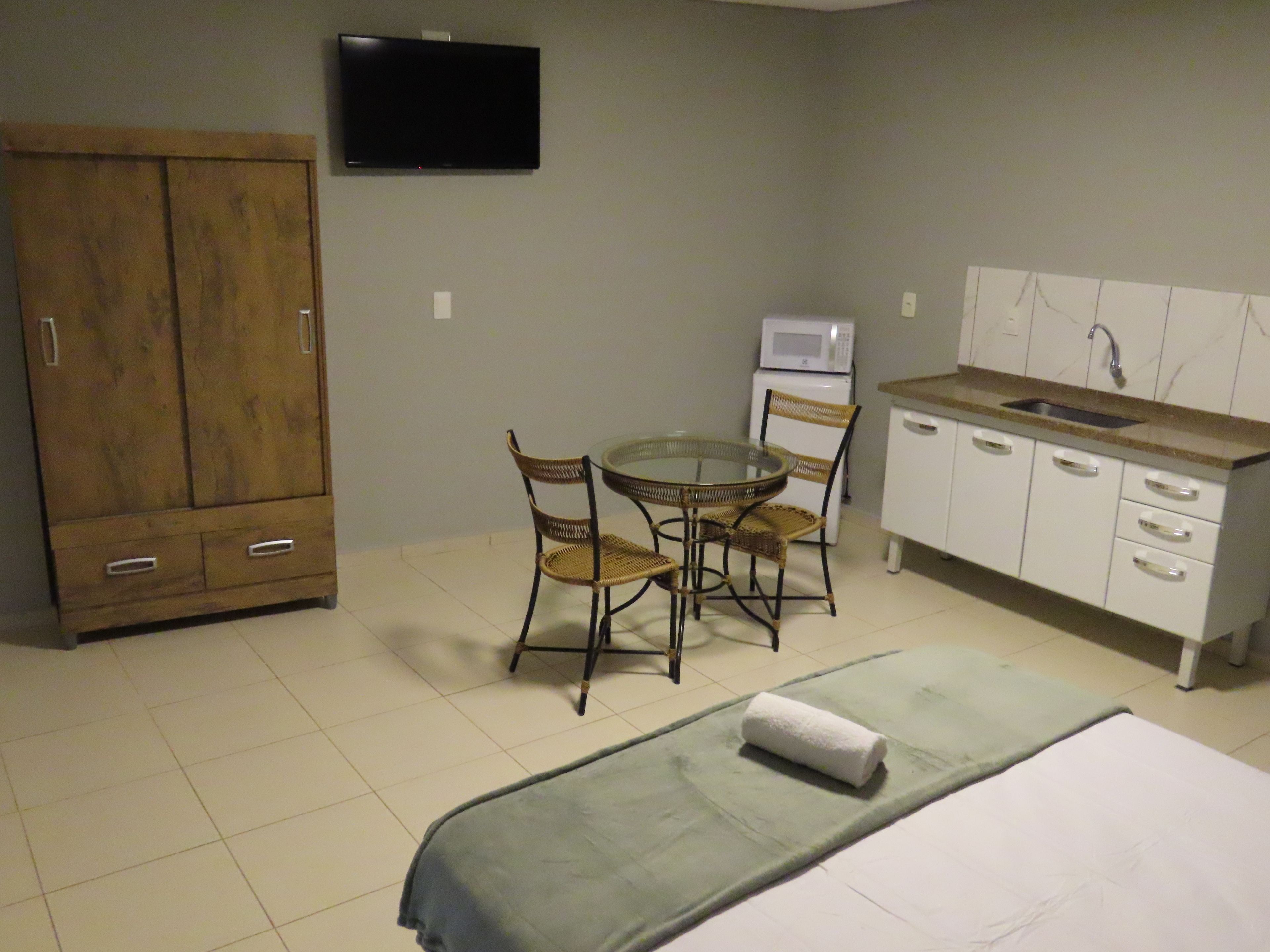 A compact room with a bed, a dining table with chairs, a kitchenette, and a TV on the wall.