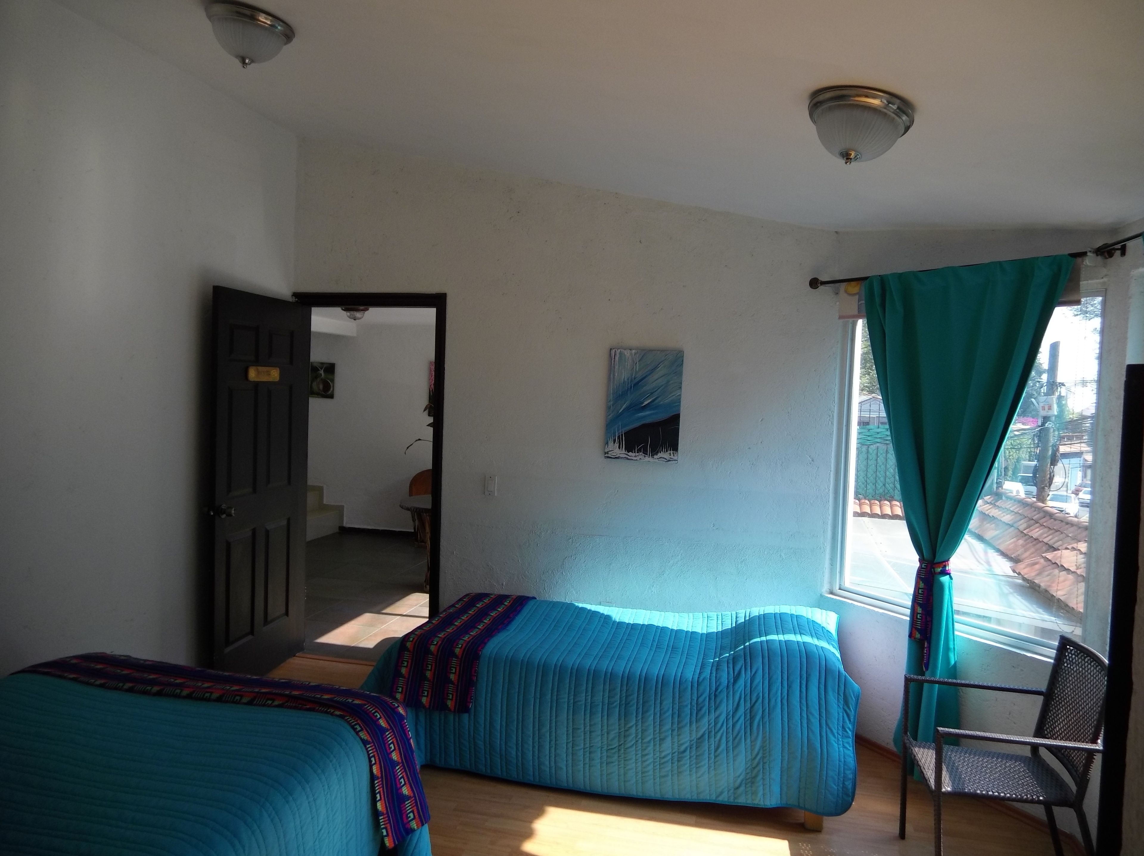 A room with two beds, a chair, a painting, and a view through a window with curtains.