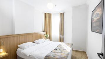 Habitació d'hotel amb un panell de capçalera de fusta, un llit amb roba de llit blanca, una tauleta de nit amb una làmpada, una finestra amb cortines i un quadre amb marc a la paret.