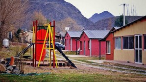 Un parque infantil con un tobogán rojo y una escalera amarilla, rodeado de edificios rojos con techos de metal, con montañas al fondo.