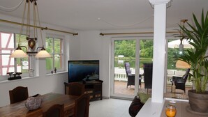 Zona pranzo con TV, mobili in legno e vista su un lago attraverso ampie finestre.