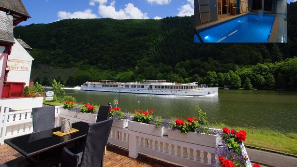 Un ristorante sul fiume con posti a sedere all'aperto, vista sul fiume, una barca ormeggiata nelle vicinanze e una collinata boscosa sullo sfondo.