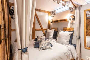 A bedroom with a wooden headboard, a bed with white bedding and blue pillows, a bedside table with a lamp, and a window with curtains. - La Grange Ungersheim - Gîte (Ungersheim)