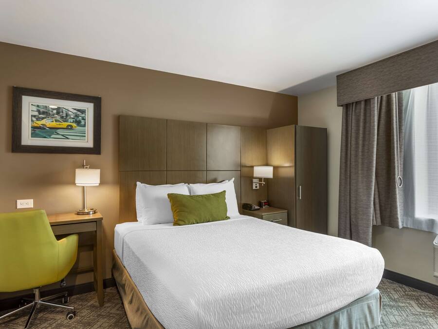 A hotel room with a large bed, a desk with a chair, a nightstand, a lamp, a framed picture on the wall, and a window with curtains.