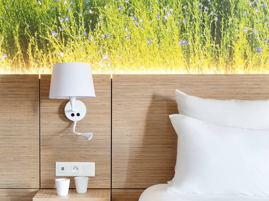 Un dormitorio con cabecera de madera, una lámpara de noche blanca, un estante pequeño con dos tazas blancas y vistas a un campo lleno de flores.