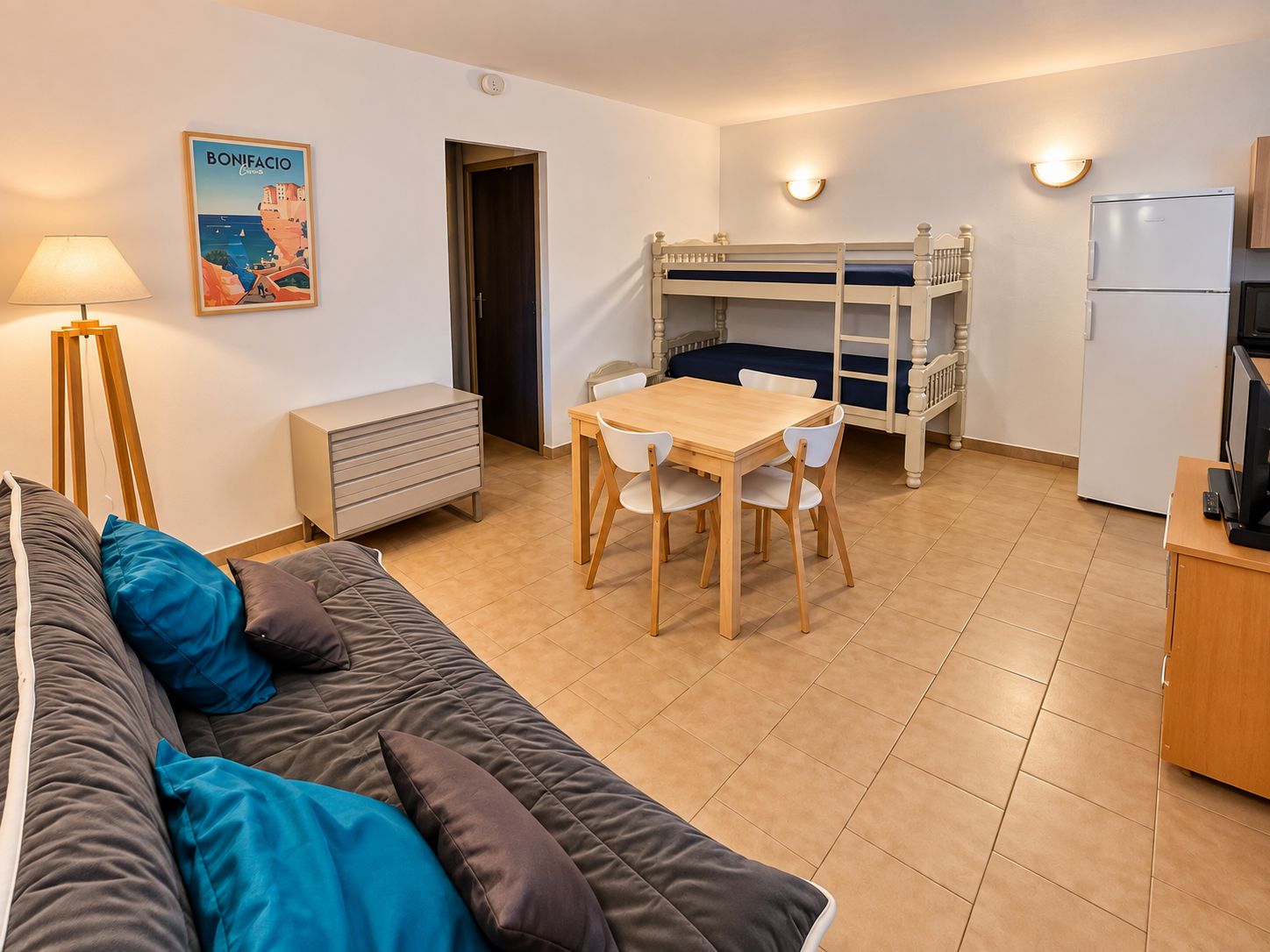 Un espacio habitable compacto con una litera, una mesa de comedor con sillas, un sofá con cojines, un escritorio con una computadora y un refrigerador.