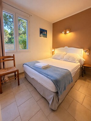 A bedroom with a bed, a chair, a nightstand, and a window with a view of trees. - Ground-floor apartment with a stunning view of the sea, garden, and mountains (Calcatoggio)