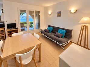 A modern living room with a sofa, a dining table, and chairs. There is a television on a stand, a lamp, and a picture on the wall. - Ground-floor apartment with a stunning view of the sea, garden, and mountains (Calcatoggio)