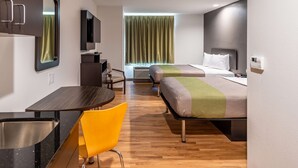 A modern hotel room with a wooden floor, a desk with a chair, a bed with a green and white blanket, a TV, and a window with curtains. - Studio 6 Lake Charles, LA (Lake Charles)