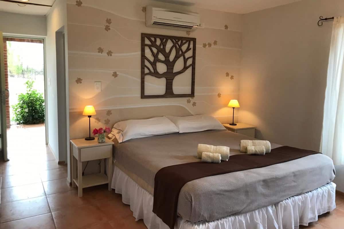 Habitación de hotel con cama, dos lámparas de noche, aire acondicionado de pared y una obra de arte con motivos de un árbol.