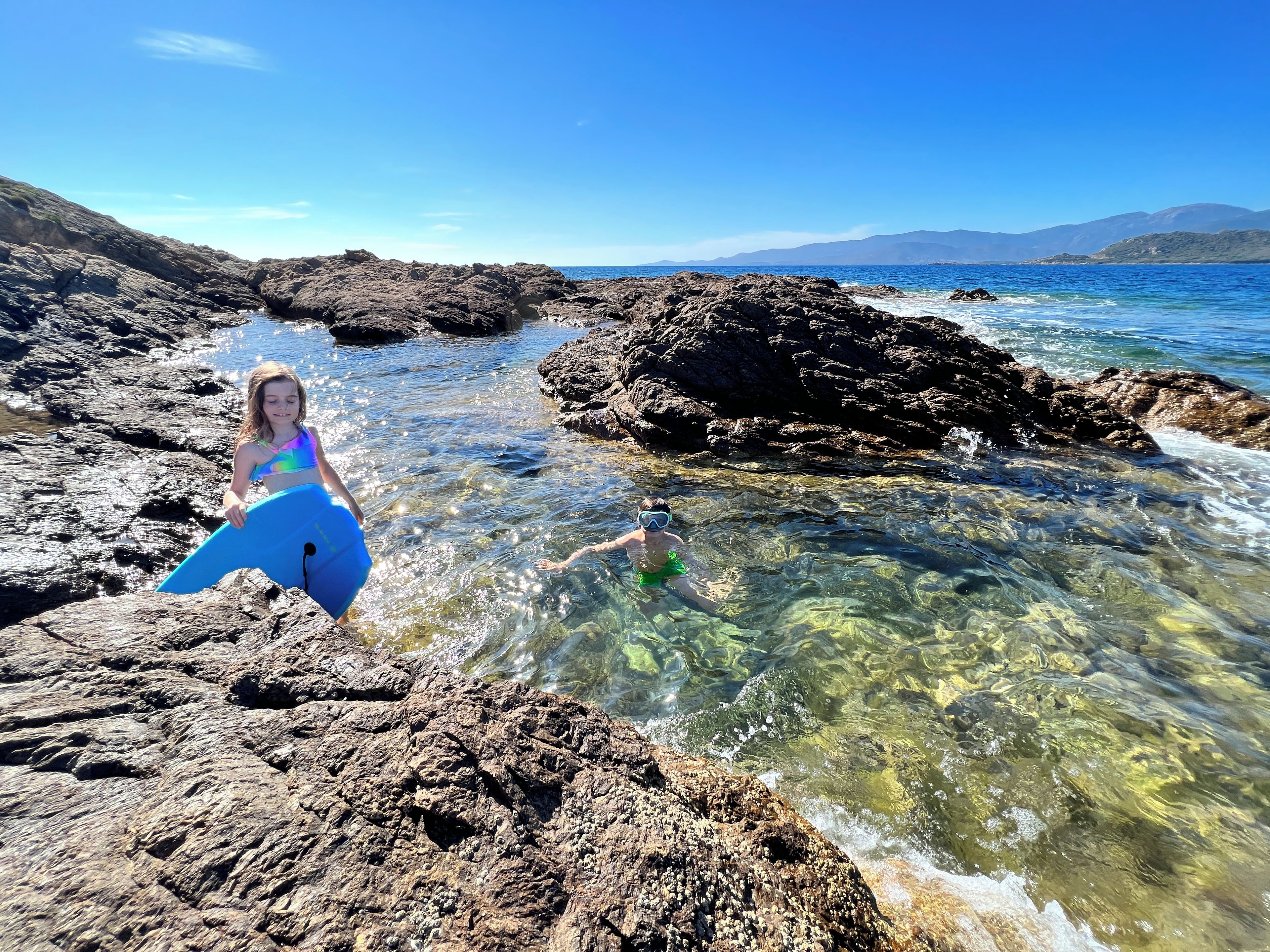 Una playa rocosa con aguas cristalinas y montañas al fondo.