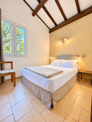 A bedroom with a bed, a chair, a nightstand, and a window with a view of trees. - Shaded apartment with views of the sea, garden, and mountains, on the upper floor (Calcatoggio)