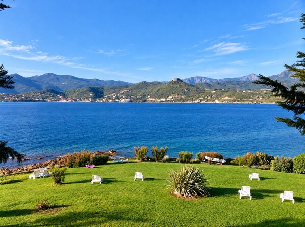 Un paisaje costero con una zona verde, sillas de descanso blancas y vistas al mar y a las montañas.